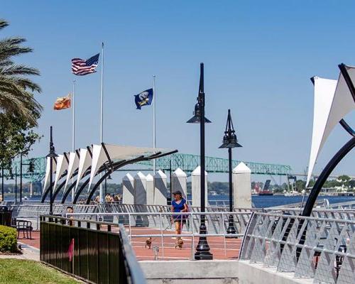 Photo of Jacksonville's Southbank Riverwalk during a sunny day. Brick sidewalk alongside the river with steel guardrails. Canopies overhang the sidewalk providing shade.